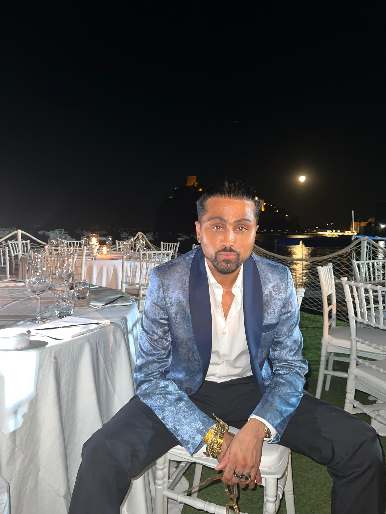 Samuel Sohebi seated at an evening event during the Ischia Global Film & Music Festival, photographed after receiving the Ischia People Award 2022, wearing a tailored evening look by the waterfront setting.